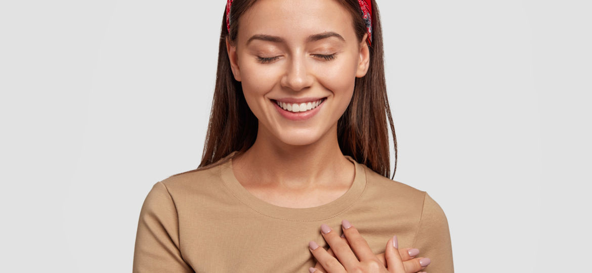 Honest kind hearted woman keeps both palms on chest, expresses kindness, keeps eyes shut, dressed in casual outfit, isolated over white background. People, feeling and body language concept.